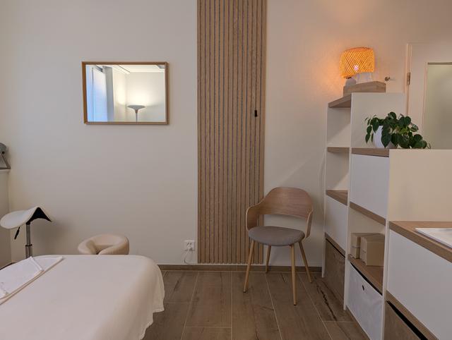 Massageraum in der Praxis origin Massage Oerlikon Bahnhof mit Liege, Wandspiegel, Holzlamellenwand und offenem Regal mit Pflanzen und dekorativer Lampe.